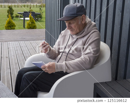 Senior male in casual attire, wearing baseball cap, writing notes while sitting relaxed in white chair on sunny home porch 125116612