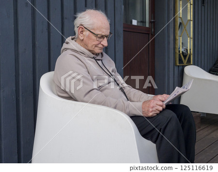Retired elderly male relaxing in garden chair, reading printed news, enjoying tranquil outdoor solitude during sunlit afternoon retirement moment Retired elderly male relaxing in garden chair, reading printed news, enjoying tranquil outdoor solitude during sunlit afternoon retirement moment 125116619