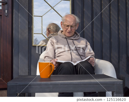 Senior man relaxing in lush garden, holding bright orange mug, reading book while savoring peaceful afternoon moment with warm beverage Senior man relaxing in lush garden, holding bright orange mug, reading book while savoring peaceful afternoon moment with warm beverage 125116620