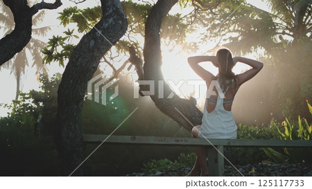 Young woman is enjoying the fresh air in a tropical forest at sunset, she is stretching her arms while sitting on a wooden fence 125117733
