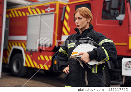 Heavy truck behind. Holding protective helmet. Woman firefighter in uniform is at work in department Heavy truck behind. Holding protective helmet. Woman firefighter in uniform is at work in department 125117735