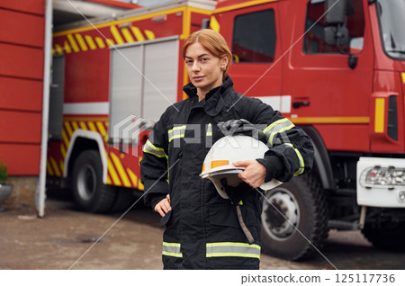Heavy truck behind. Holding protective helmet. Woman firefighter in uniform is at work in department 125117736