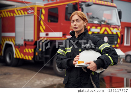 Heavy truck behind. Holding protective helmet. Woman firefighter in uniform is at work in department Heavy truck behind. Holding protective helmet. Woman firefighter in uniform is at work in department 125117737