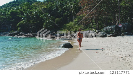 A lone figure strolls along a sandy beach, surrounded by lush palm trees and a tranquil sea. The sun casts a warm glow over the serene landscape, inviting relaxation. A lone figure strolls along a sandy beach, surrounded by lush palm trees and a tranquil sea. The sun casts a warm glow over the serene landscape, inviting relaxation. 125117771
