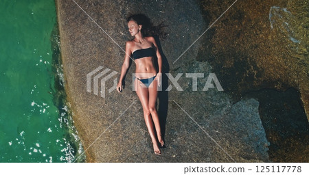 A woman in a bikini lies on a large rock by clear turquoise water, soaking up the sun. The tranquil environment reflects a perfect day for relaxation and leisure by the sea. 125117778