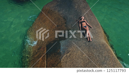 A woman relaxes under the warm sun on a smooth rock formation, enjoying the tranquility of the turquoise waters around her. This serene moment captures a perfect day at a tropical location. A woman relaxes under the warm sun on a smooth rock formation, enjoying the tranquility of the turquoise waters around her. This serene moment captures a perfect day at a tropical location. 125117793