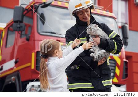 Cute scottish fold cat is in the hands. Firefighter woman in uniform is with a little girl Cute scottish fold cat is in the hands. Firefighter woman in uniform is with a little girl 125117819