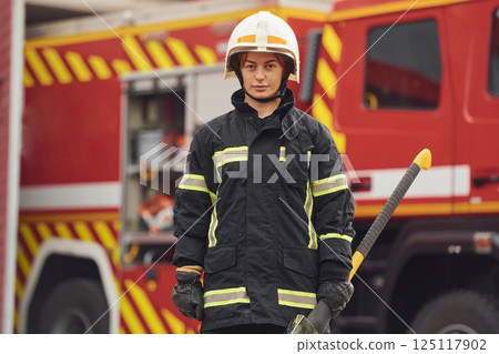 Standing with axe in hands against truck. Woman firefighter in uniform is at work in department Standing with axe in hands against truck. Woman firefighter in uniform is at work in department 125117902