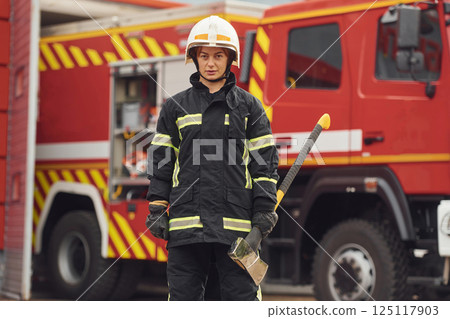 Standing with axe in hands against truck. Woman firefighter in uniform is at work in department Standing with axe in hands against truck. Woman firefighter in uniform is at work in department 125117903