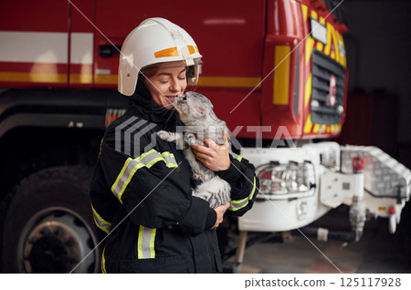 Standing against big truck and holding beautiful cat. Woman firefighter in uniform is at work in department Standing against big truck and holding beautiful cat. Woman firefighter in uniform is at work in department 125117928
