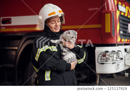 Cat with grey wool is in the hands of employee. Woman firefighter in uniform is at work in department 125117929