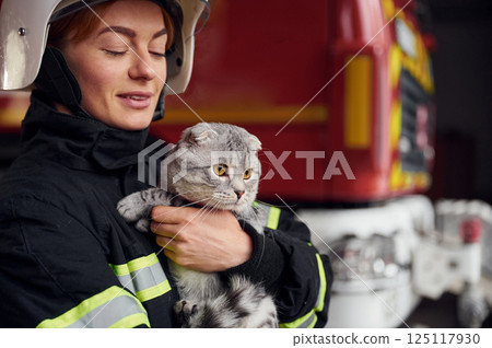 Cat with grey wool is in the hands of employee. Woman firefighter in uniform is at work in department Cat with grey wool is in the hands of employee. Woman firefighter in uniform is at work in department 125117930