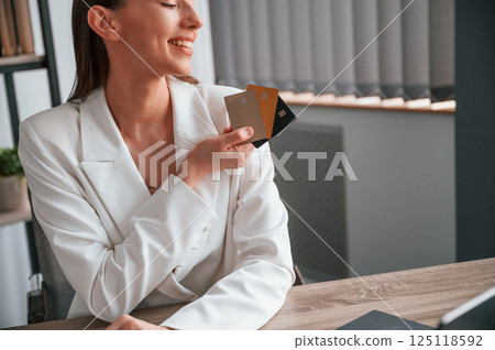 Sitting and holding three credit cards. Young beautiful woman in formal clothes is working at office 125118592