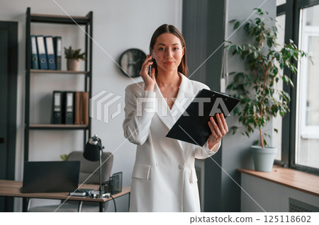 With notepad and smartphone in hands. Young beautiful woman in formal clothes is working at office 125118602