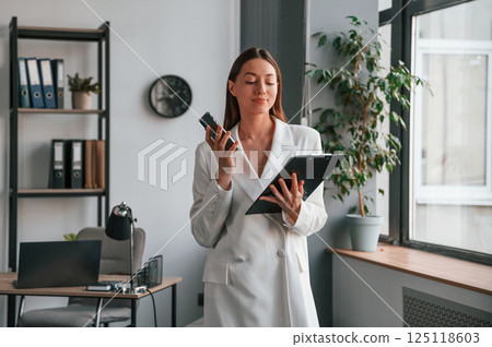 With notepad and smartphone in hands. Young beautiful woman in formal clothes is working at office With notepad and smartphone in hands. Young beautiful woman in formal clothes is working at office 125118603