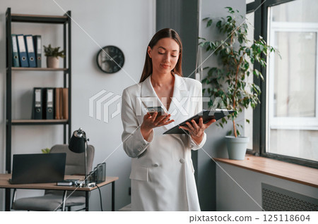 With notepad and smartphone in hands. Young beautiful woman in formal clothes is working at office With notepad and smartphone in hands. Young beautiful woman in formal clothes is working at office 125118604