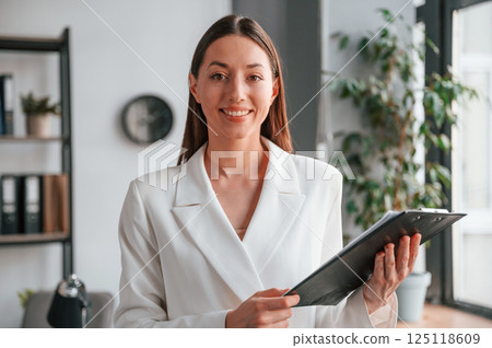 Standing and holding notepad. Young beautiful woman in formal clothes is working at office Standing and holding notepad. Young beautiful woman in formal clothes is working at office 125118609