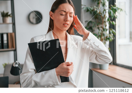 Holding notepad and having headache. Young beautiful woman in formal clothes is working at office Holding notepad and having headache. Young beautiful woman in formal clothes is working at office 125118612