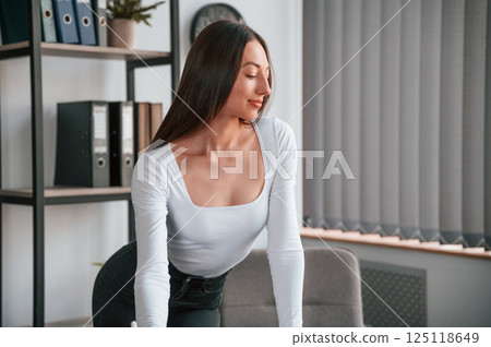 Standing by the table. Young beautiful woman in white clothes is in the office 125118649