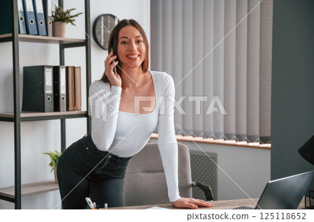Standing and talking by phone. Young beautiful woman in white clothes is in the office 125118652