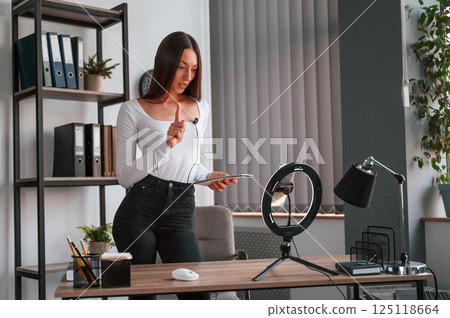 Smartphone is on the stand, recording. Young beautiful woman in white clothes is in the office 125118664