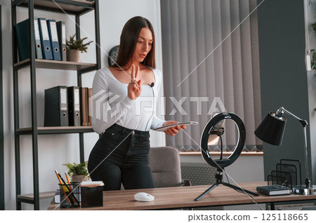 Smartphone is on the stand, recording. Young beautiful woman in white clothes is in the office 125118665