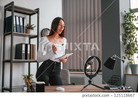 Smartphone is on the stand, recording. Young beautiful woman in white clothes is in the office 125118667