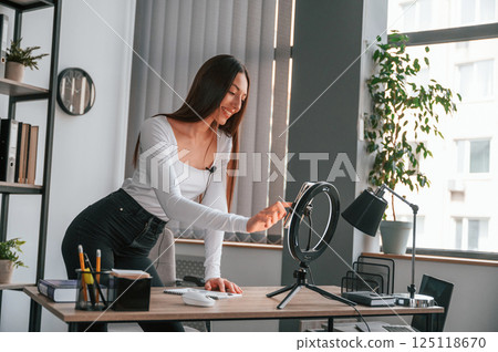 Setting up the smartphone for recording. Young beautiful woman in white clothes is in the office 125118670