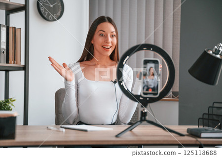 Smartphone that on the stand is recording. Online master class. Young beautiful woman in white clothes is in the office Smartphone that on the stand is recording. Online master class. Young beautiful woman in white clothes is in the office 125118688