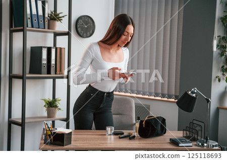 Standing and using laptop. Young beautiful woman in white clothes is in the office 125118693