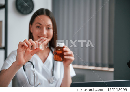 Holding pills in hands. Young beautiful female doctor in white coat is in the office Holding pills in hands. Young beautiful female doctor in white coat is in the office 125118699