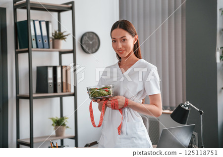 Holding salad and measuring tape. Conception of diet. Young beautiful woman in white clothes is in the office Holding salad and measuring tape. Conception of diet. Young beautiful woman in white clothes is in the office 125118751