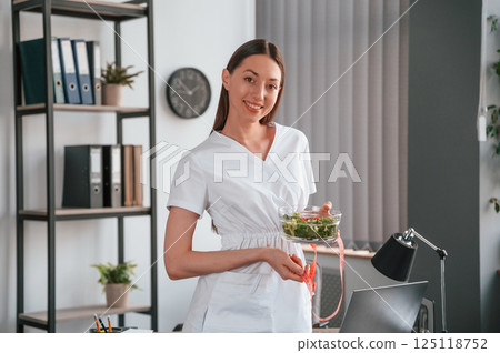 Holding salad and measuring tape. Conception of diet. Young beautiful woman in white clothes is in the office 125118752