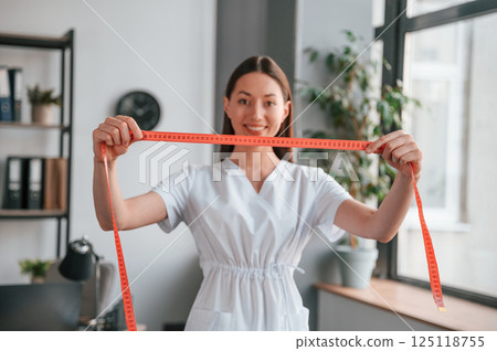 Standing with measuring tape. Young beautiful woman in white clothes is in the office Standing with measuring tape. Young beautiful woman in white clothes is in the office 125118755