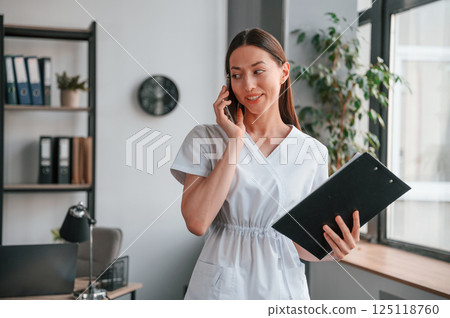 Standing, talking by phone and holding notepad. Young beautiful woman in white clothes 125118760
