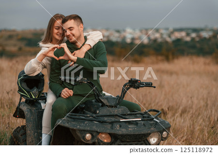 Making love shape gesture by hands. Beautiful young couple is with quad bike outdoors on the field 125118972
