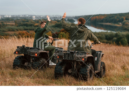 Beautiful landscape, giving high five. Two men are on the quad bikes outdoors on the field Beautiful landscape, giving high five. Two men are on the quad bikes outdoors on the field 125119084