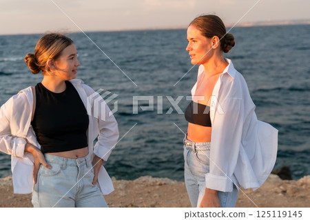 Women Beach Sunset Conversation - Two women stand on a beach at sunset, looking at each other and talking. 125119145