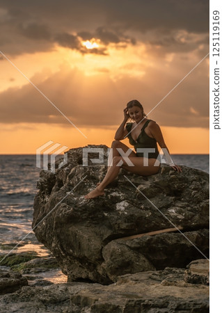 Woman Sunset Beach - Silhouette of woman sitting on a rock at sunset with the ocean behind her. Woman Sunset Beach - Silhouette of woman sitting on a rock at sunset with the ocean behind her. 125119169