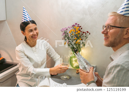 Couple Birthday Celebration Kitchen: Happy couple toasts with wine, flowers, party hats in cozy kitchen celebrating birthday. Couple Birthday Celebration Kitchen: Happy couple toasts with wine, flowers, party hats in cozy kitchen celebrating birthday. 125119199