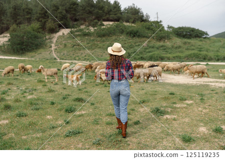 A woman wearing a straw hat walks through a field of sheep 125119235