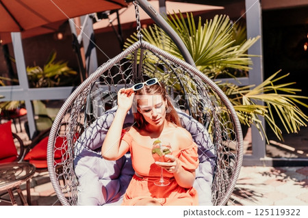 A woman in an orange dress is sitting in a hanging chair. She is holding a wine glass and smiling. The scene takes place in a restaurant with several chairs and tables. 125119322