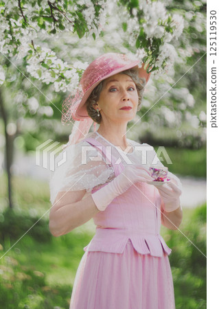 Natural adult mature woman of 60 years old in 18th century style pink hat, retro dress, gloves holds cup of tea in blossoming white apple orchard on summer day Natural adult mature woman of 60 years old in 18th century style pink hat, retro dress, gloves holds cup of tea in blossoming white apple orchard on summer day 125119530