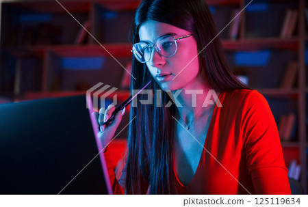 Holding stylus. Beautiful woman in glasses and red wear is sitting by the laptop in dark room with neon lighting Holding stylus. Beautiful woman in glasses and red wear is sitting by the laptop in dark room with neon lighting 125119634