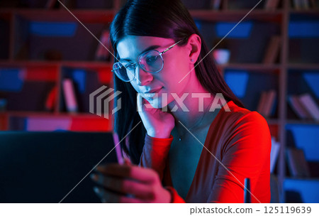 Focused on the work. Beautiful woman in glasses and red wear is sitting by the laptop in dark room with neon lighting Focused on the work. Beautiful woman in glasses and red wear is sitting by the laptop in dark room with neon lighting 125119639