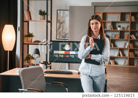 Standing against pc and holding notepad. Young female designer is in the modern office Standing against pc and holding notepad. Young female designer is in the modern office 125119815