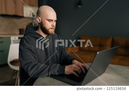 Focused programmer wearing dark hoodie typing code on laptop computer , sitting at table near bright window in contemporary urban apartment with minimalist design and soft daylight. 125119830