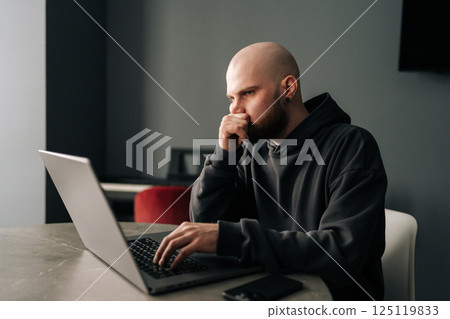 Focused freelancer young man remote working from home office, sitting at a table with laptop computer, contemplating strategies for upcoming project while reflecting on challenges and deadlines ahead Focused freelancer young man remote working from home office, sitting at a table with laptop computer, contemplating strategies for upcoming project while reflecting on challenges and deadlines ahead 125119833