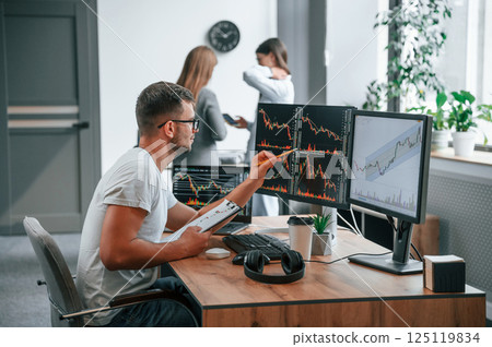 Stock broker is sitting by computer and working with exchange technology. Women is standing and talking behind 125119834