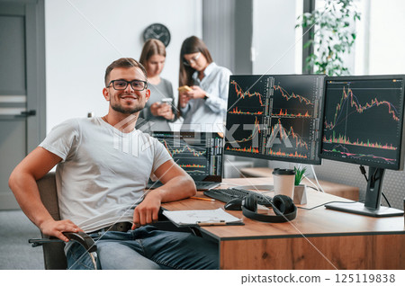 Stock broker is sitting by computer and working with exchange technology. Women is standing and talking behind Stock broker is sitting by computer and working with exchange technology. Women is standing and talking behind 125119838